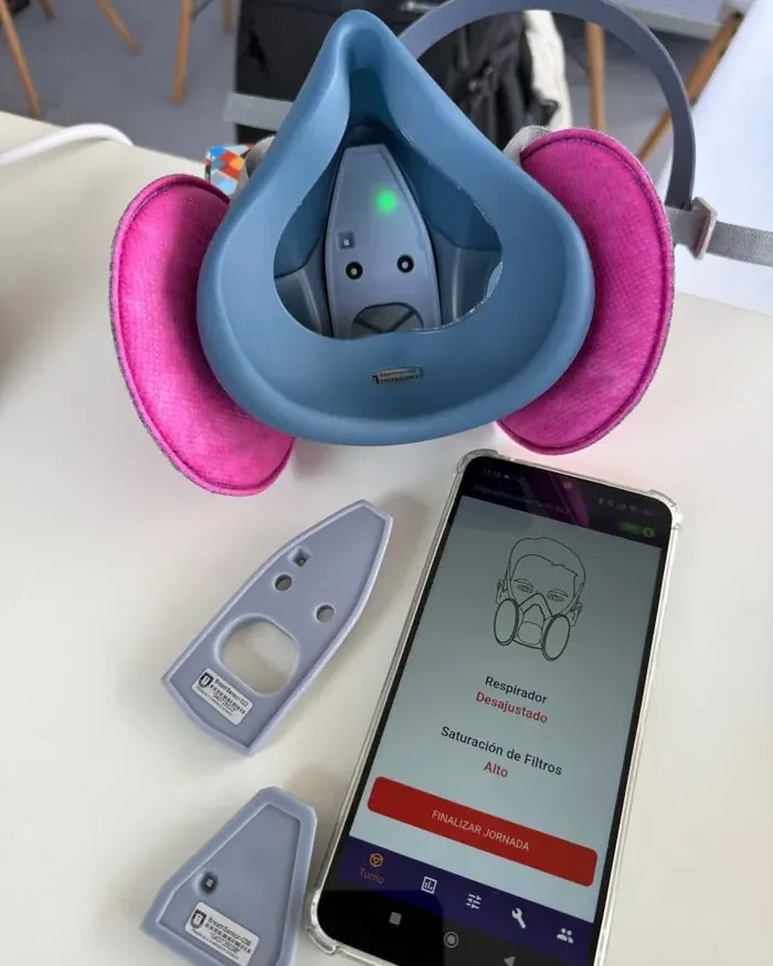 Primer plano del sensor AirWatch instalado en un respirador industrial, junto a la app móvil mostrando el estado de ajuste y saturación de filtros